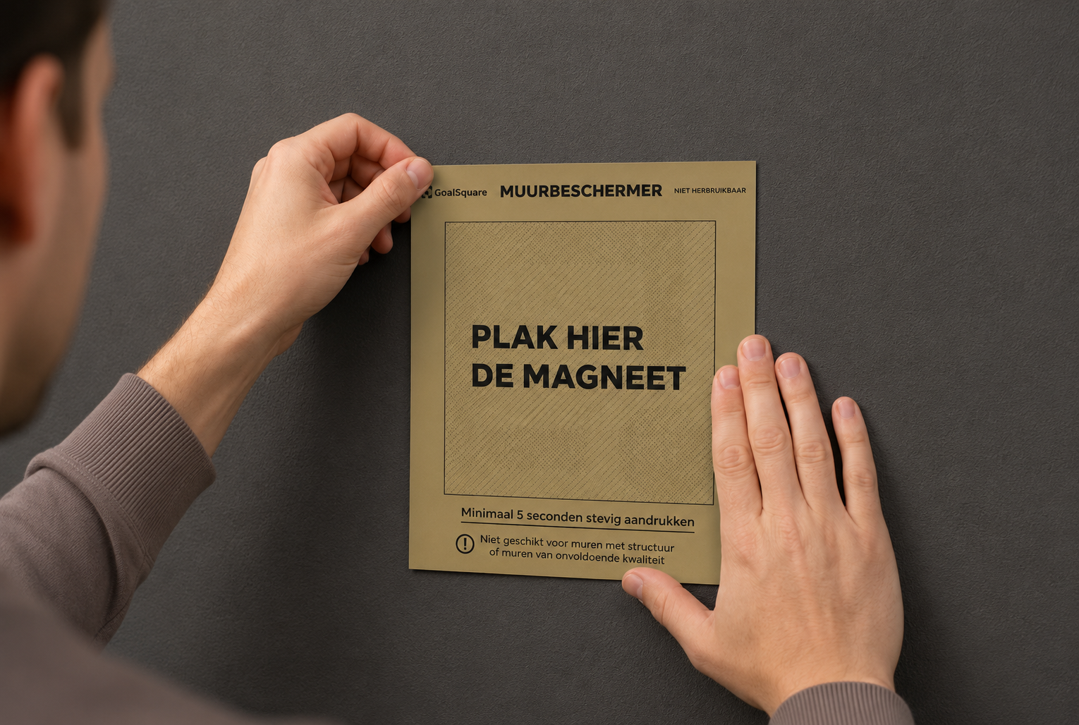 Stap 2 voor het ophangen van een GoalSquare poster: Twee handen plakken de bruingele muurbeschermer met de tekst plak hier de magneet strak op de donkere wand.